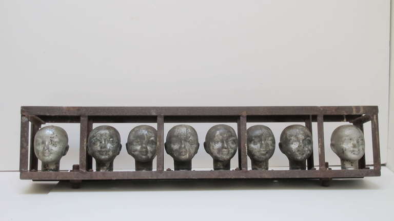 Graphic series of metal doll heads lined up in an industrial steel framing. The heads are classical in appearance and have a patina of use and silver paint.
Can be set on table shelf or easily hung on a wall.