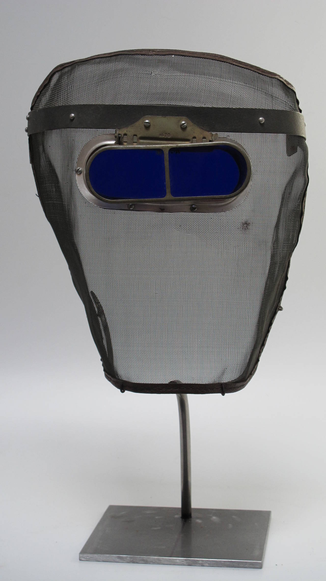 Face protection from hot cinders in tending a coal furnace, the mesh would block most particles and the blue glass would protect the eyes. Mounted as a sculpture and mask on a black metal base. The cobalt blue glass has metal framing and is hinged