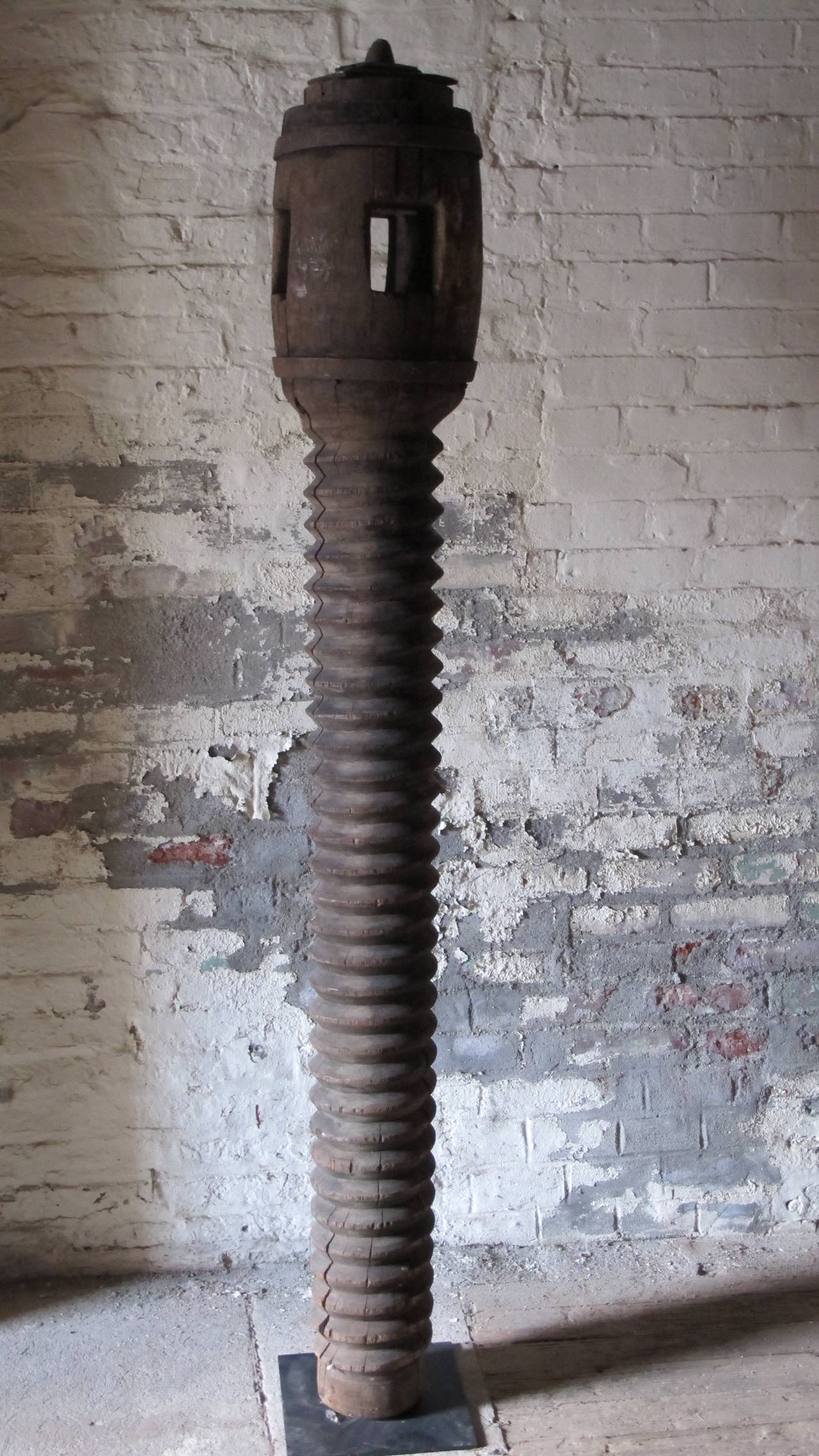 Probably made for use in making olive oil as a screw press, Most of its length is a threaded screw with a wider barrel shaped top with rectangular slots cut through. Mounted on a black metal floor stand.