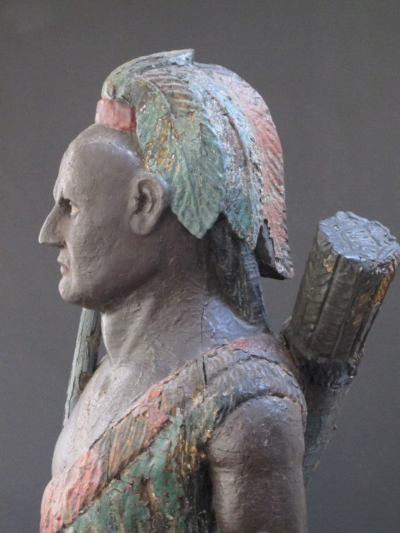 Carved wood cigar store Indian holding a bow with quiver of arrows on it's back.  The figure retains layers of old paint. The muscular Indian figure shows both strength and dignity in its portrayal.  The figure is holding tobacco leaves and is shown
