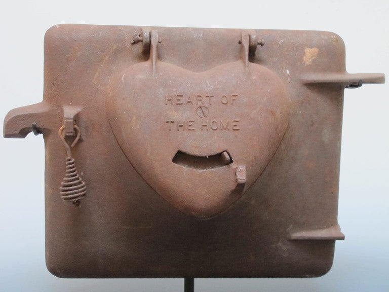The cast iron stove door has a raised  heart form that hinges upwards to look inside.  Raised lettering HEART OF THE HOME Printed on the heart with slide vent below. The hinged heart can be lifted to open revealing a heart shaped opening. Mounted