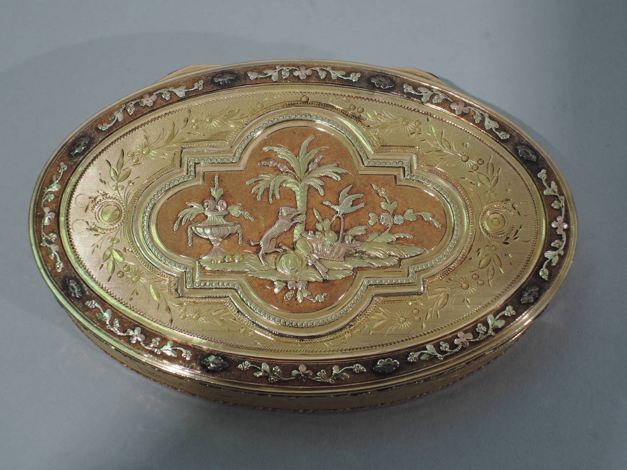 Swiss 20-karat gold snuffbox, circa 1810. Oval with hinged cover. Ornament in three-color gold. On cover are a Grecian lamp vase, wicker basket, dog, and bird. On bottom are a floral garland and baluster vase. Fine craftsmanship and rich patina.