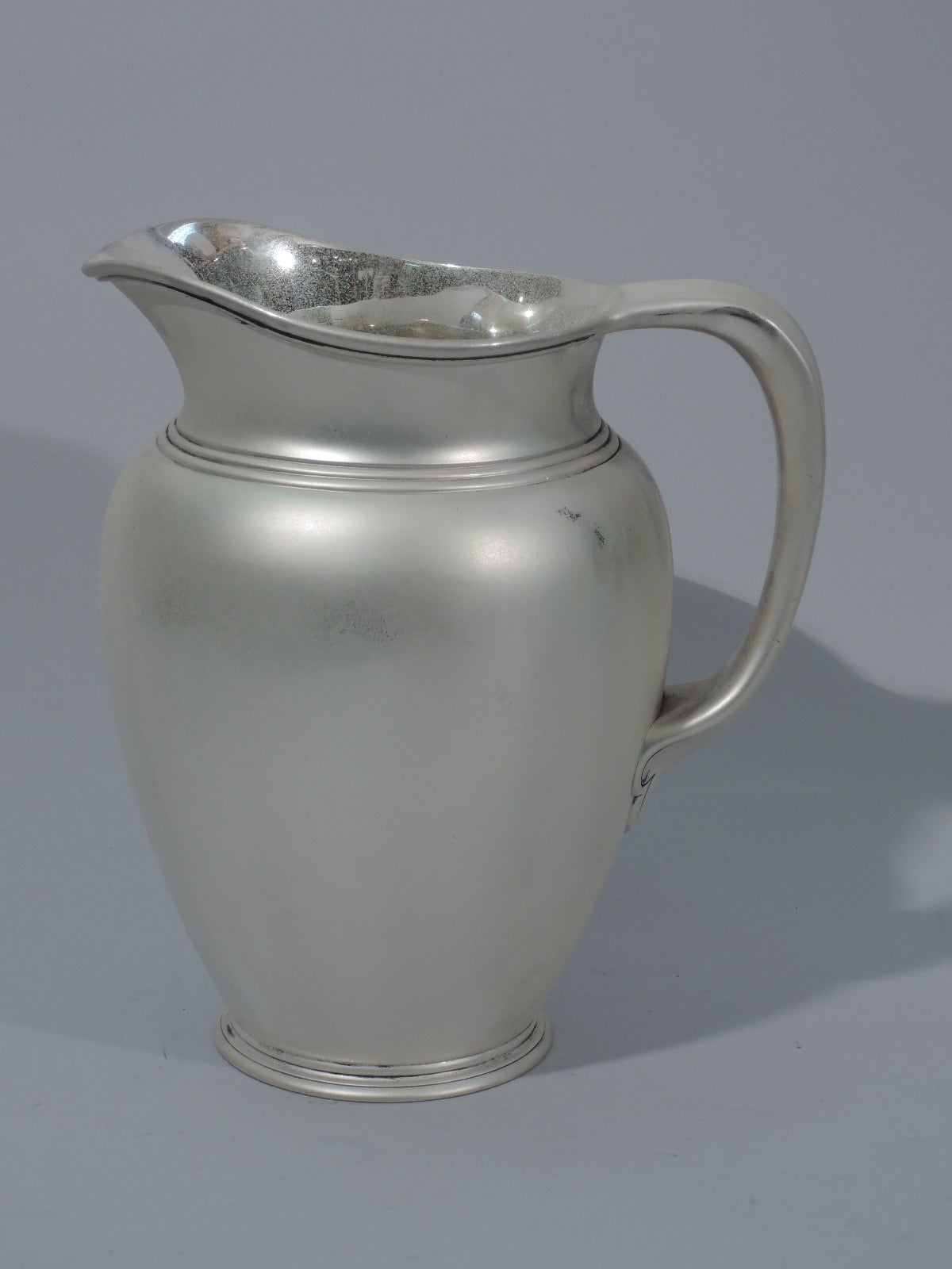 Sterling silver water pitcher. Made by Tiffany & Co. in New York, circa 1907. Ovoid body, raised foot, soft bracket handle, and molded mouth with lip spout. Chased bands at neck base. Spare and heavy. The pattern (no. 16974) was first produced in