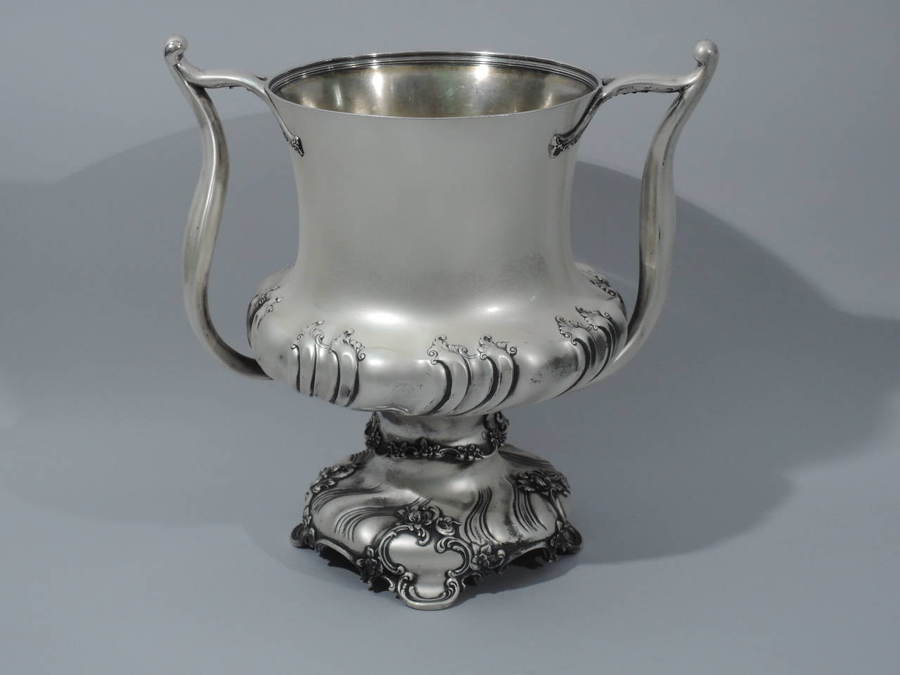 Sterling silver trophy cup. Made by Tiffany & Co. in New York, ca 1893. Squat bowl, raised foot on four C-scrolls, and flared rim. Twisted gadrooning to bowl and foot. Sinuous side handles, splayed at top with flowers and foliage. Gilt interior. A