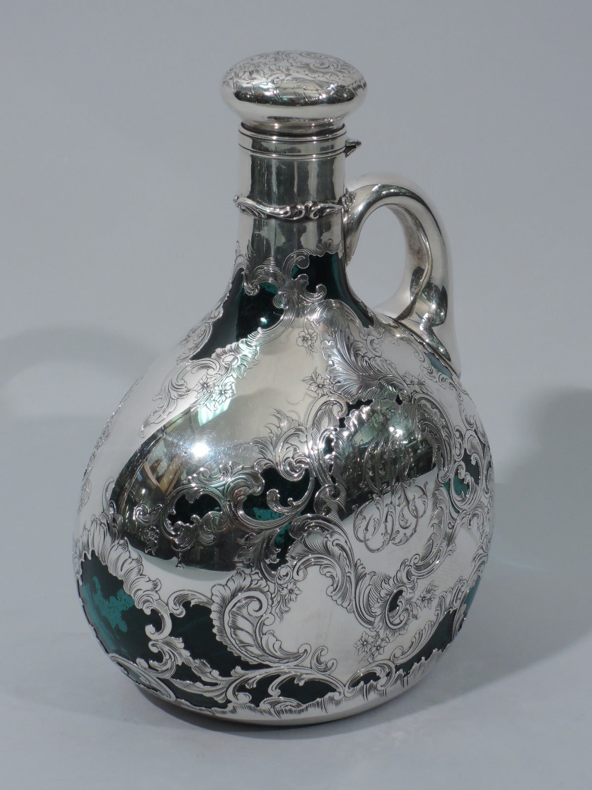 Emerald glass jug decanter with silver overlay. Made by Gorham in Providence, circa 1890. Globular body, and cylindrical neck. Engraved and chased flowers, foliage, and scrolls, with peeps of rich green. Bun cover is hinged and cork lined with same