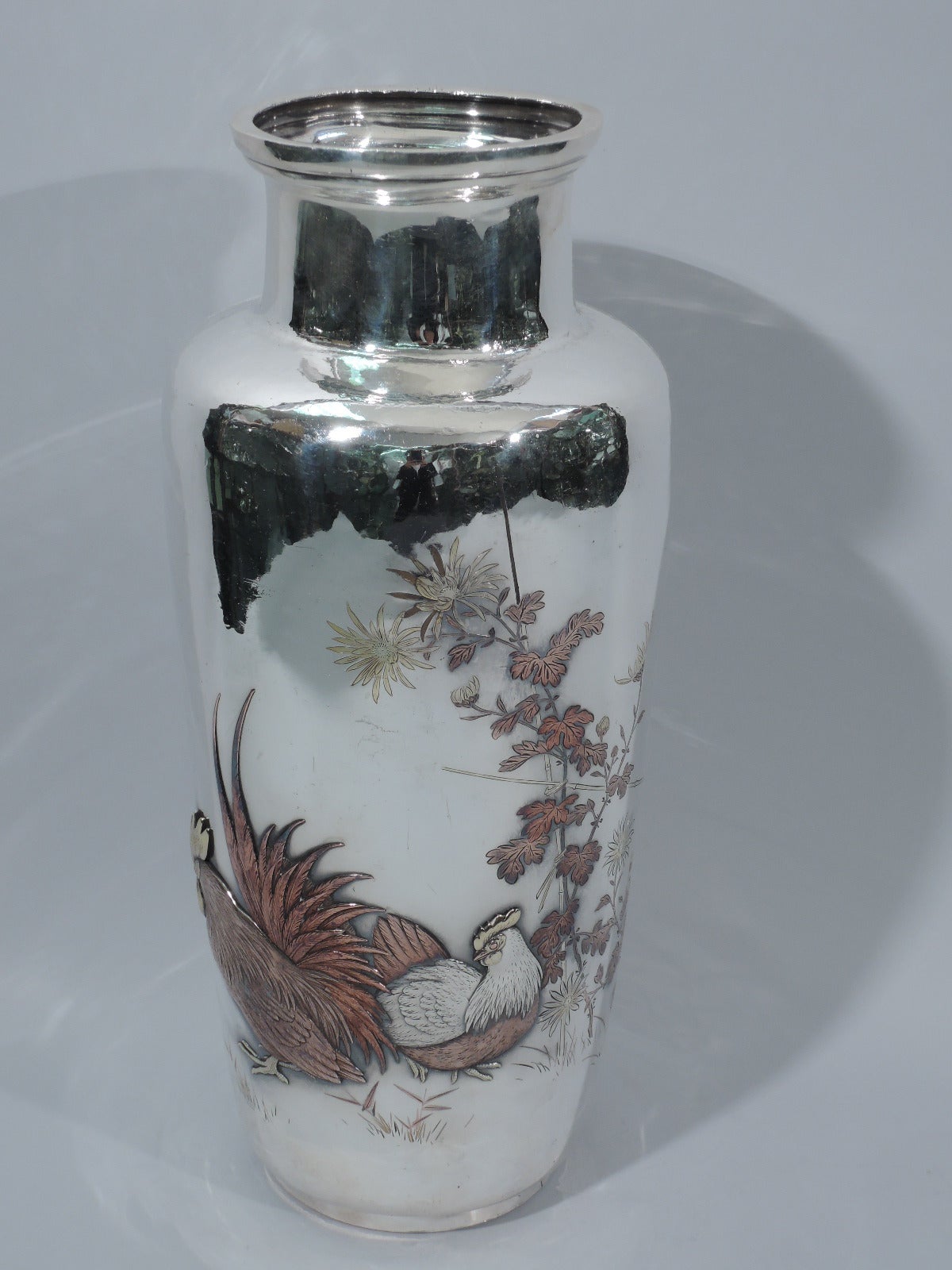 Large silver vase with mixed-metal farmyard scene. Made by Kobayashi in Tokyo. Baluster with straight and short neck, flat rim, and foot ring. Applied to body: a rooster and hen warily face-down an advancing chick. Inlaid bamboo, flowers, and grass.
