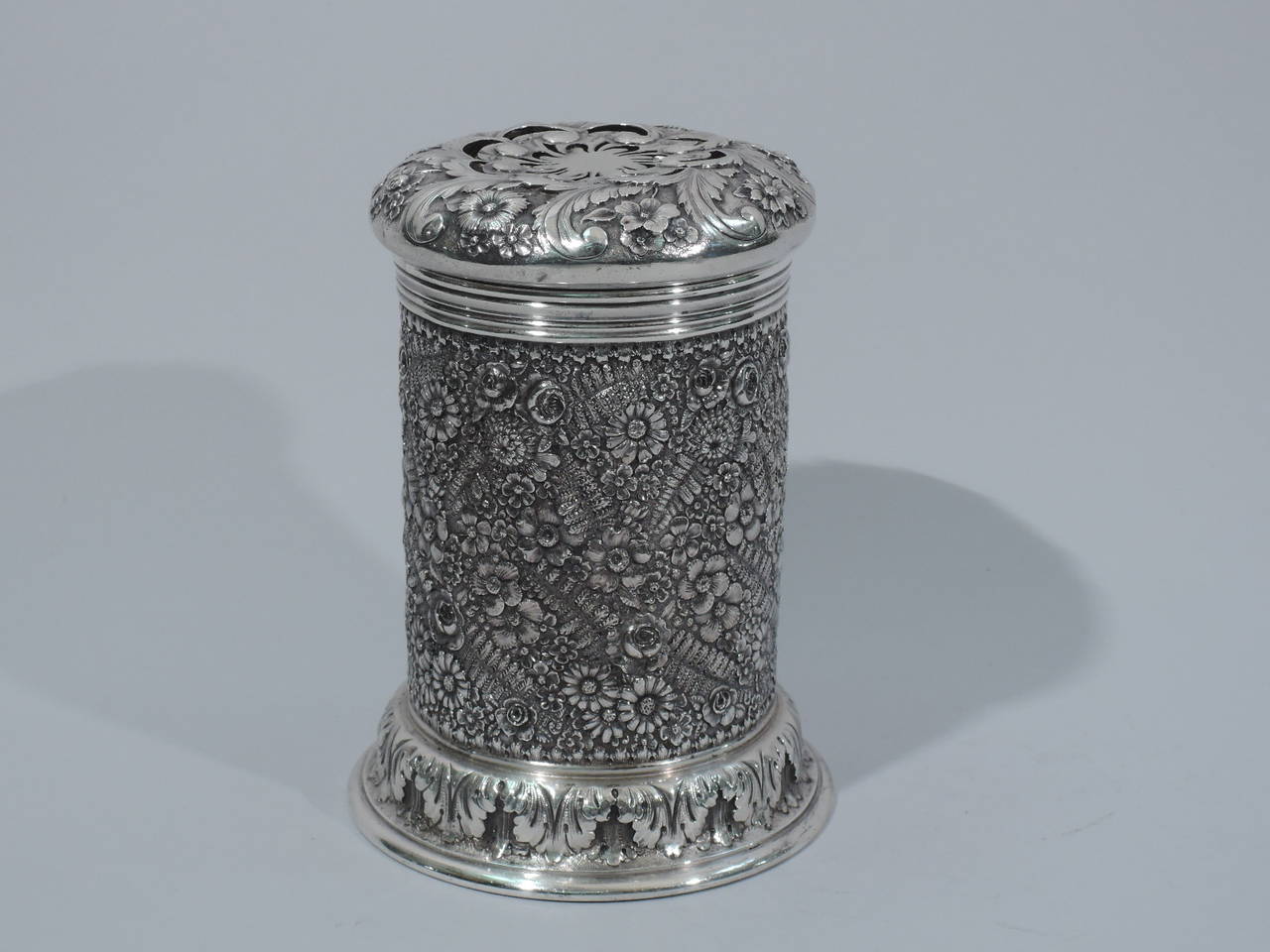 Unusual sterling silver shaker. Made by Tiffany & Co. in New York, ca. 1887. Cylindrical with spread foot and bun cover. Dense repousse with flowers and ferns. Acanthus leaf border to foot. Reeded rim. Cover has scrolled piercing bordered by