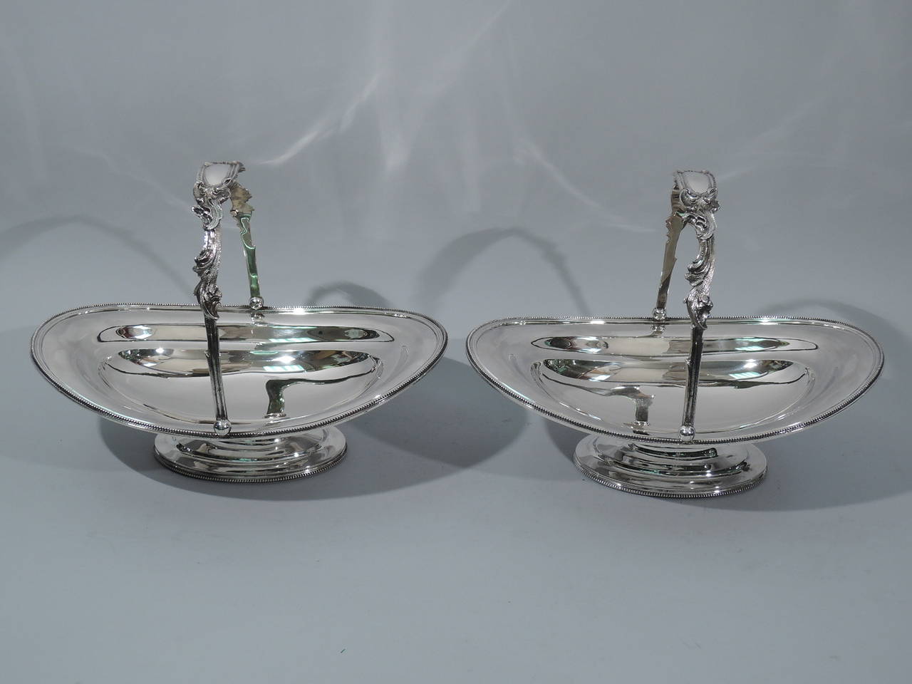 Pair of sterling silver baskets. Retailed by Tiffany & Co. at 550 Broadway, circa 1854-1855. Each shallow and oval bowl on stepped oval foot. Beaded rims. Chased and scrolled swing handle with tubular cartouche (vacant). Rare early Tiffany. Hallmark