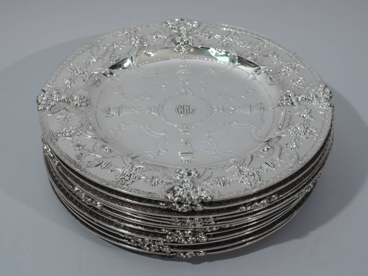 Set of 12 Renaissance-Revival sterling silver service plates. Made by historic Tiffany & Co. in New York. Each: circular with deep well. Raised ornament includes strapwork and rosette frames and borders. Also, covered vases, and bunches of fruits
