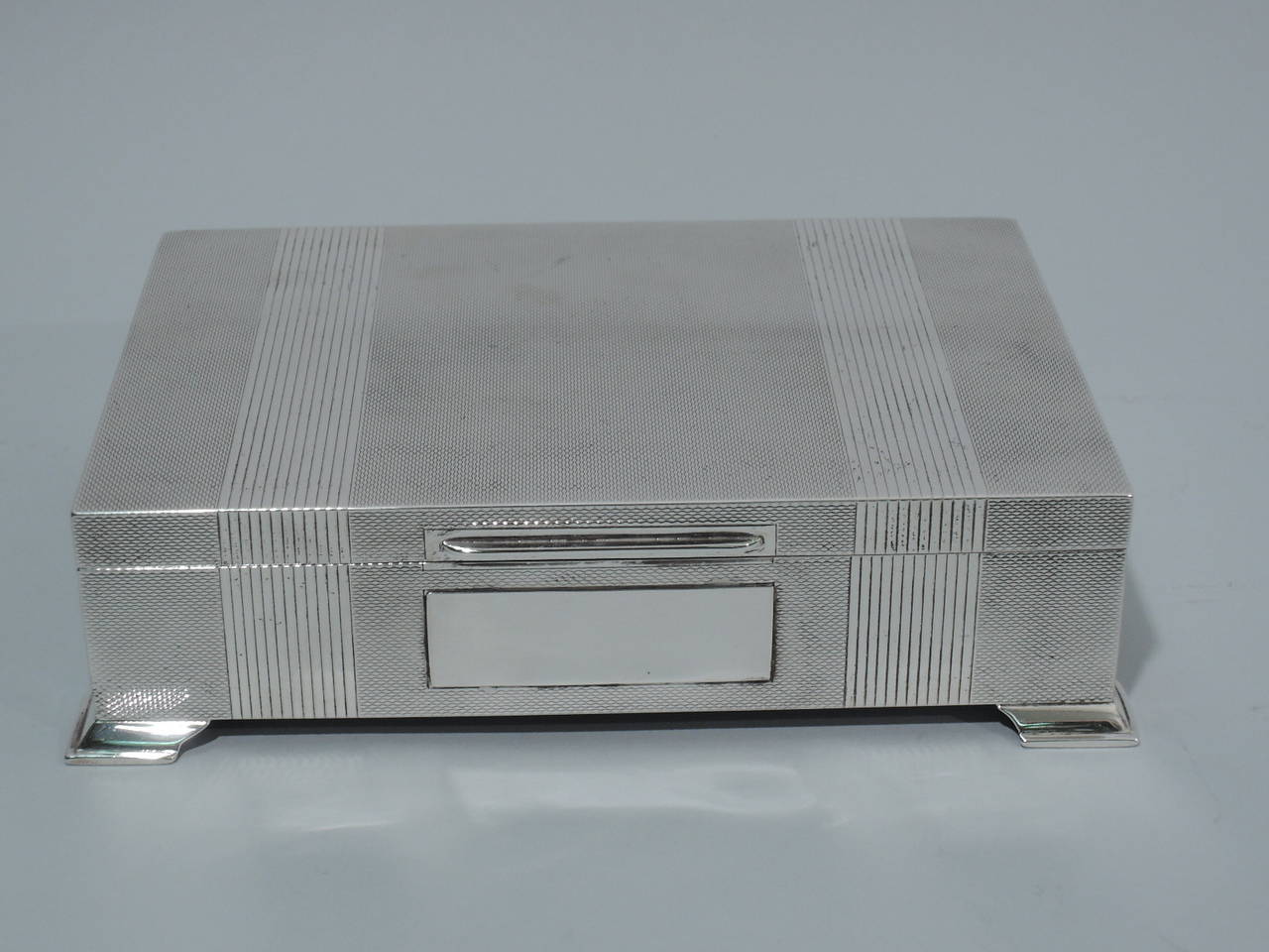 Elizabeth II sterling silver desk box. Made by Harman Bros. in Birmingham in 1958. Rectangular with spread bracket feet at corners. Cover is hinged and flat with tab. Allover engine-turned pattern. On cover are two reeded bands that wrap around to