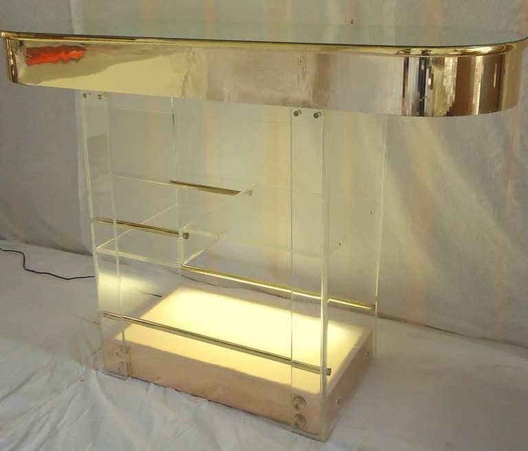 Elegant Brass and Lucite bar manufactured by Hill Industries. The mirrored top is inset. The top is bordered by an etched border of brass topped by a brass coated 1/2 round. 
The base lights up from an inline switch and there is an 8 bottle storage