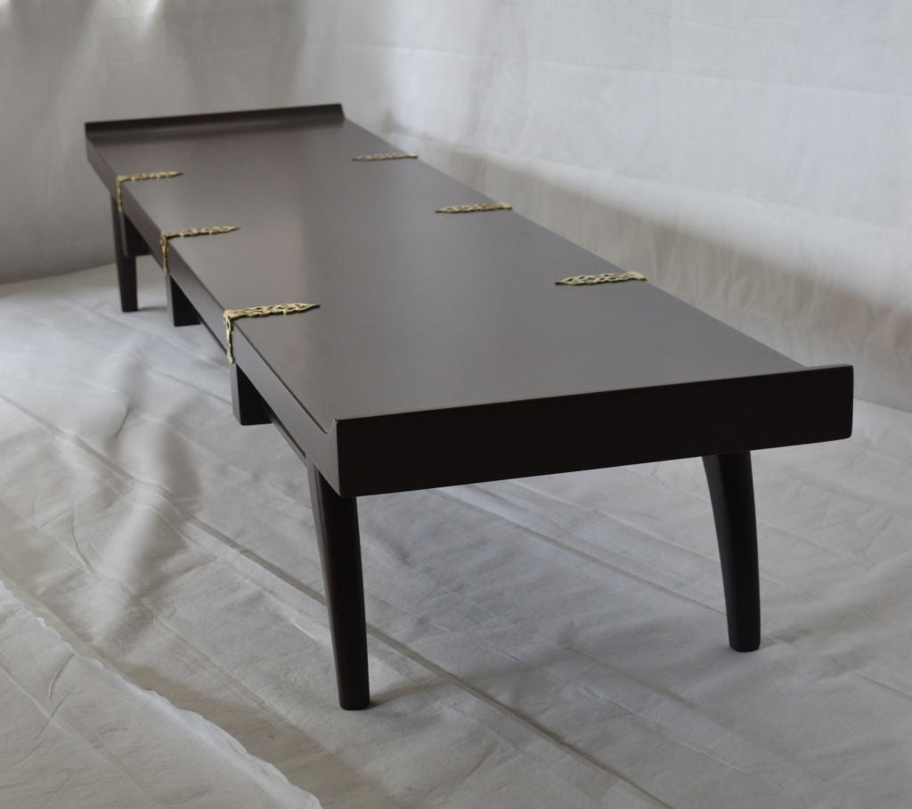 A mahogany cocktail table with with brass filigree straps, carved block details and elegantly arched legs designed by Edmond Spence and manufactured in Mexico in the mid-1950s. 

This table is part of  Edmund Spence's Industria Mueblara line of