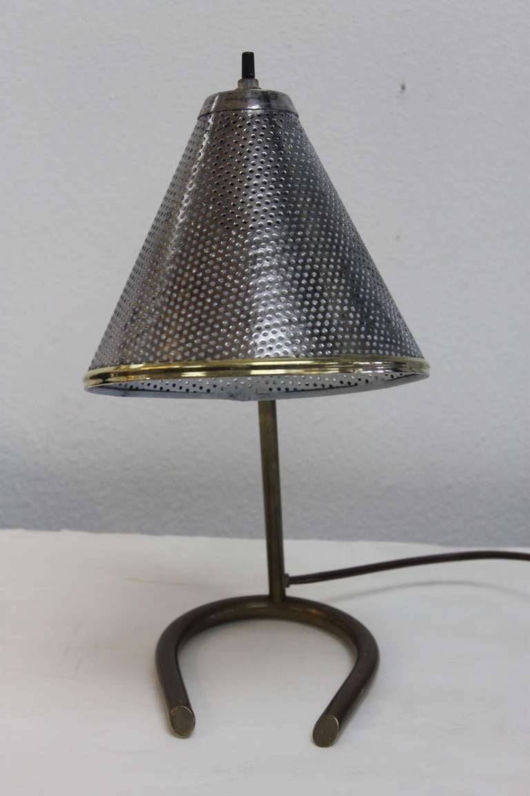 Small electric table lamp. Lamp with a brass base and a perforated cone shade, nickel plated with brass trim. The shade is adjustable by means of a small brass wing-nut. On/off turn switch is located at the top of the cone. Overall height is 13”.