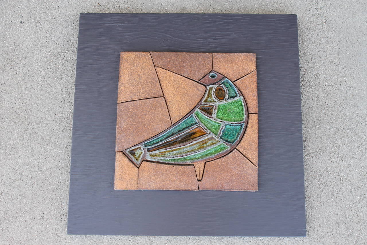 Circa 1960's earthenware tile mosaic depicting an abstract bird form with pooled glass sections in blue, green and gold. The mosaic is affixed to its original painted wood backing (repainted). The wood panel measures 20.75