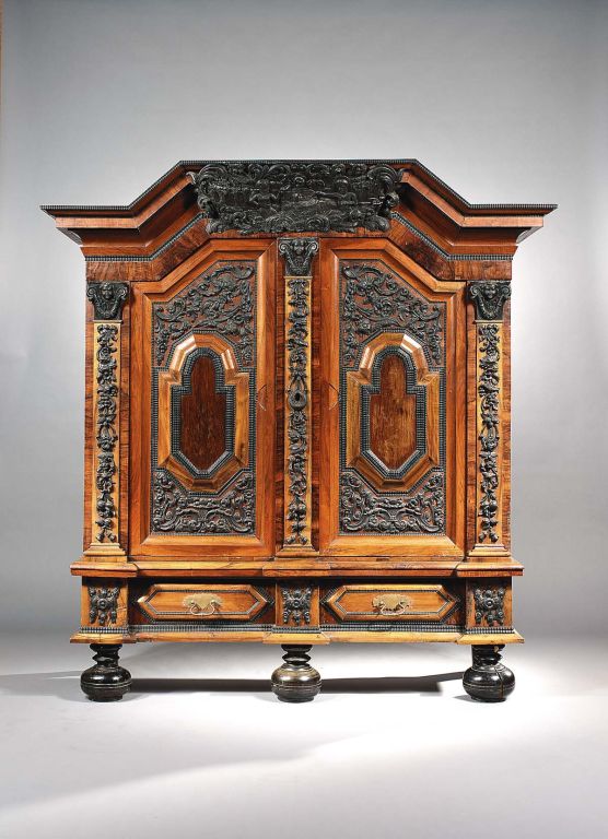 A German Baroque Walnut and Ebonised Cupboard from the Saxe-Coburg collection. The arched cornice rises over a profusely carved cartouche. The two doors feature raised panels of Walnut amidst intricate Ebonised carvings, the doors are divided by