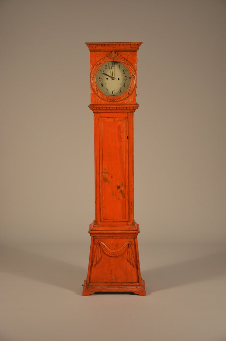 Danish tall clock scraped back to the old red surface. The clock bonnet has a wreath surrounding the face and the face has Arabic numerals. Round corners flank the raised panel door like a country column. Dental molding above and below the face. And
