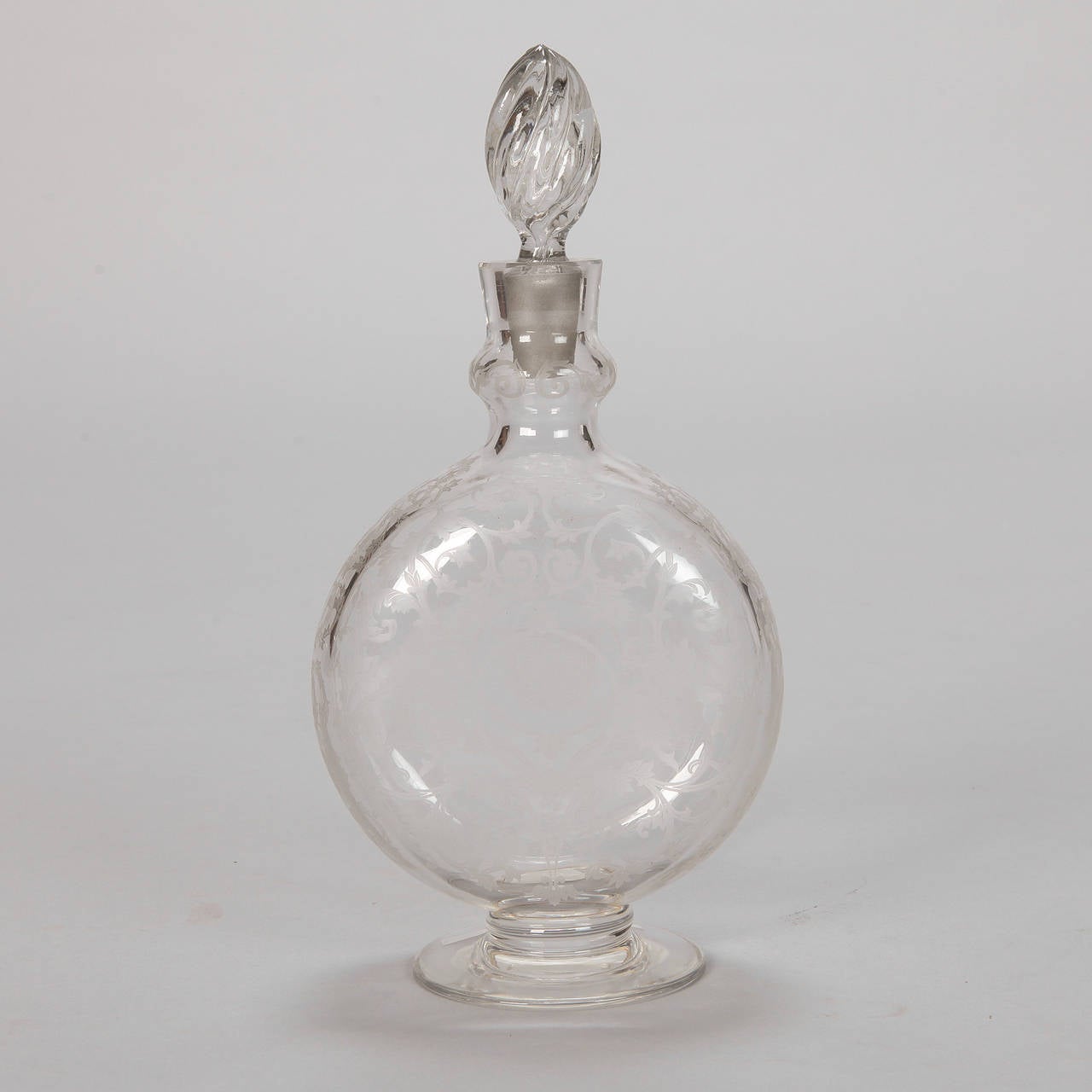 Circa 1930s signed Baccarat decanter of clear, etched glass with pedestal base, round, disk-shape body decorated with etched vines and flowers and a tall, twisted flame-shape stopper. Etched signature on the bottom.