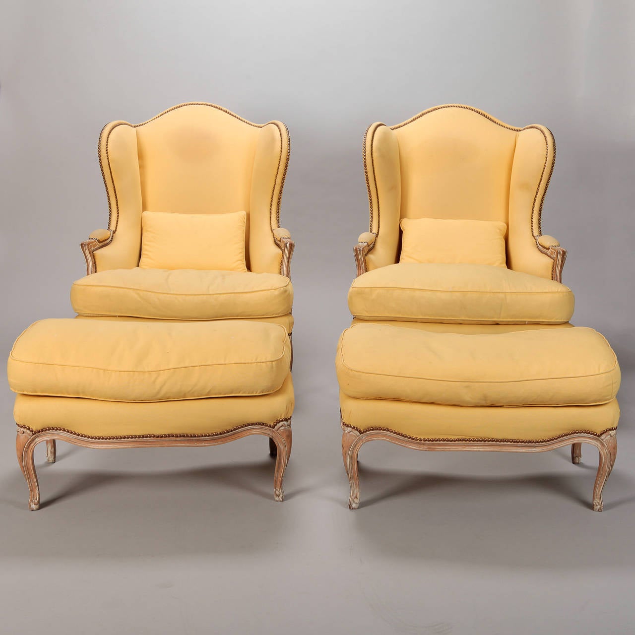 Pair circa 1920s down filled wing chairs found in France with coordinating ottomans. Details include carved wood frames, padded arms and decorative brass nail heads. Yellow fabric upholstery shows some wear and discoloration.  Structurally, the