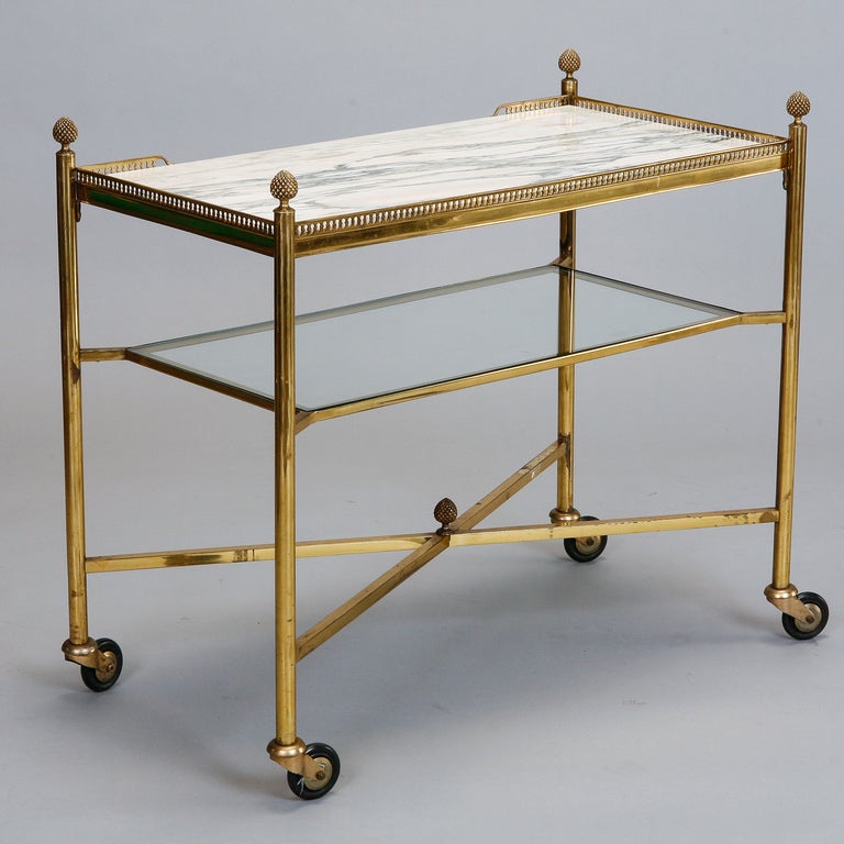 his circa 1940s brass framed trolley table has white and gray marble table top with a lower glass shelf, X-base stretcher, wheels and pineapple finials. 