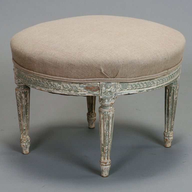 Circa 1920s round French stool has a carved frame with tapered, reeded legs and painted antique white finish. Newly upholstered in a natural linen. 