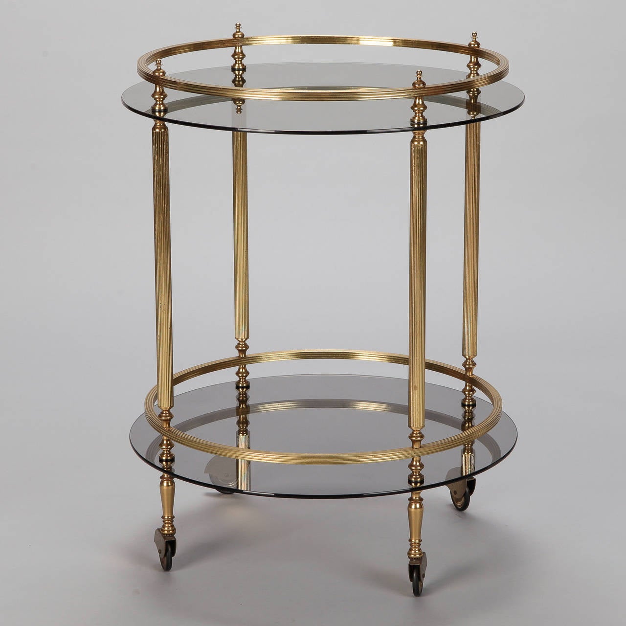 Circa 1960s two tier drinks trolley / server has a brass frame with round, smoked glass tops. Brass casters, brass legs and rails are reeded and topped with finials for a timeless, neoclassical look.