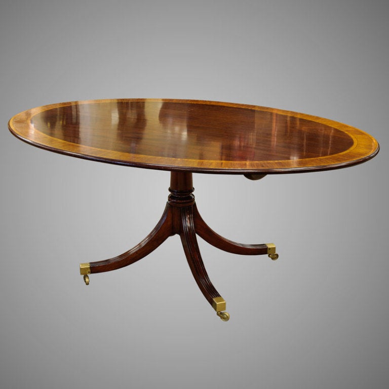 Circa 1920s English mahogany tilt top table has an oval top with contrasting banded edge. The base has four reeded legs with brass caps and casters.