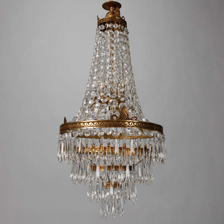 This circa 1920 French empire style chandelier has a decorative brass frame with crested detail supports four graduated tiers of crystal pendants at the bottom and draped beading at the top. Ceiling canopy is 4.5