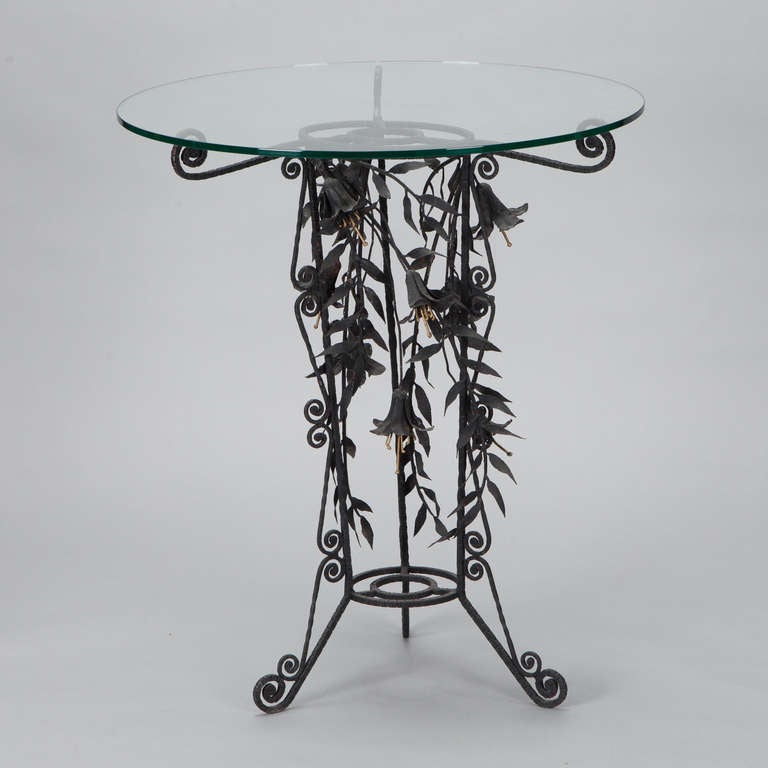 Circa 1930s side table features a black iron tri-foot base with intricately rendered tole lilies and scrolled support arms. Shown here with a clear glass top which can be changed for a larger glass top or for a top of another material. Suitable for