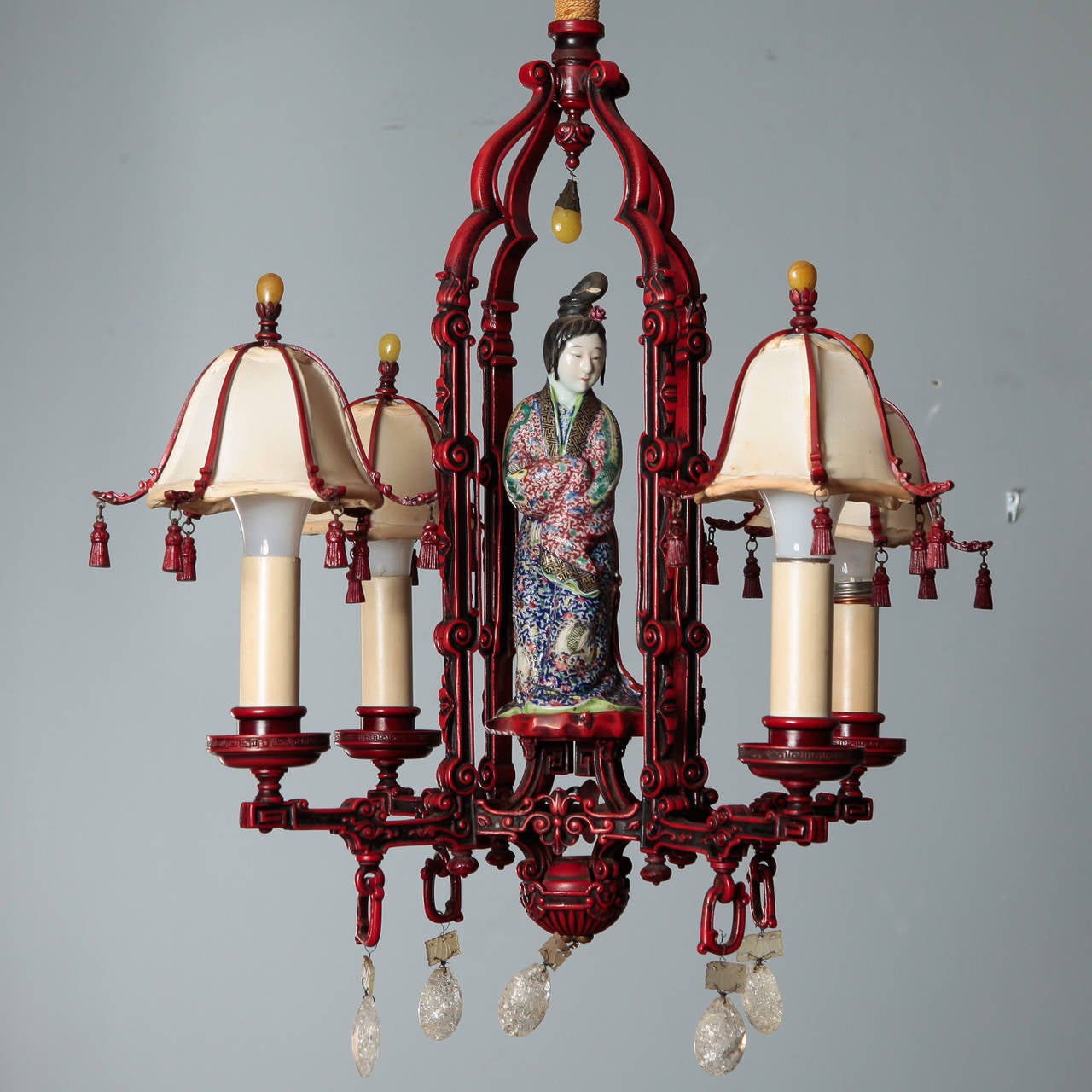 Circa 1920s Chinoserie chandelier with iron base in red painted finish with candle style lights, original silk shades with metal tassels, a central porcelain figure and rock crystal pendants. Updated wiring. Sconces in this style also available.