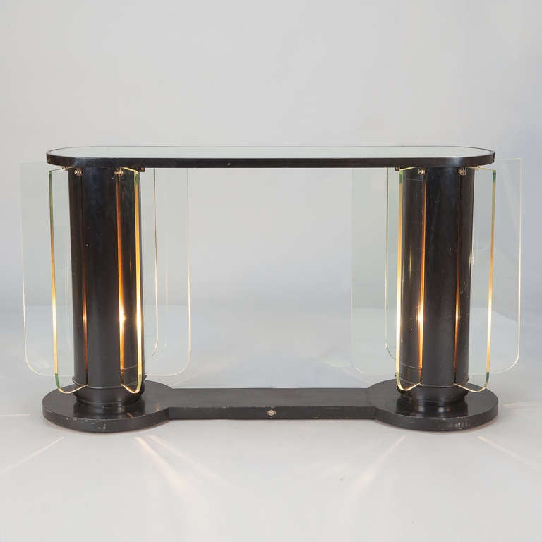 Circa 1930s French Art Deco console with an ebonised finish, oval mirrored glass table top and column shaped supports with internal lights and glass insert panels. Internal lights create a dramatic illumination of the radiating glass fin-like