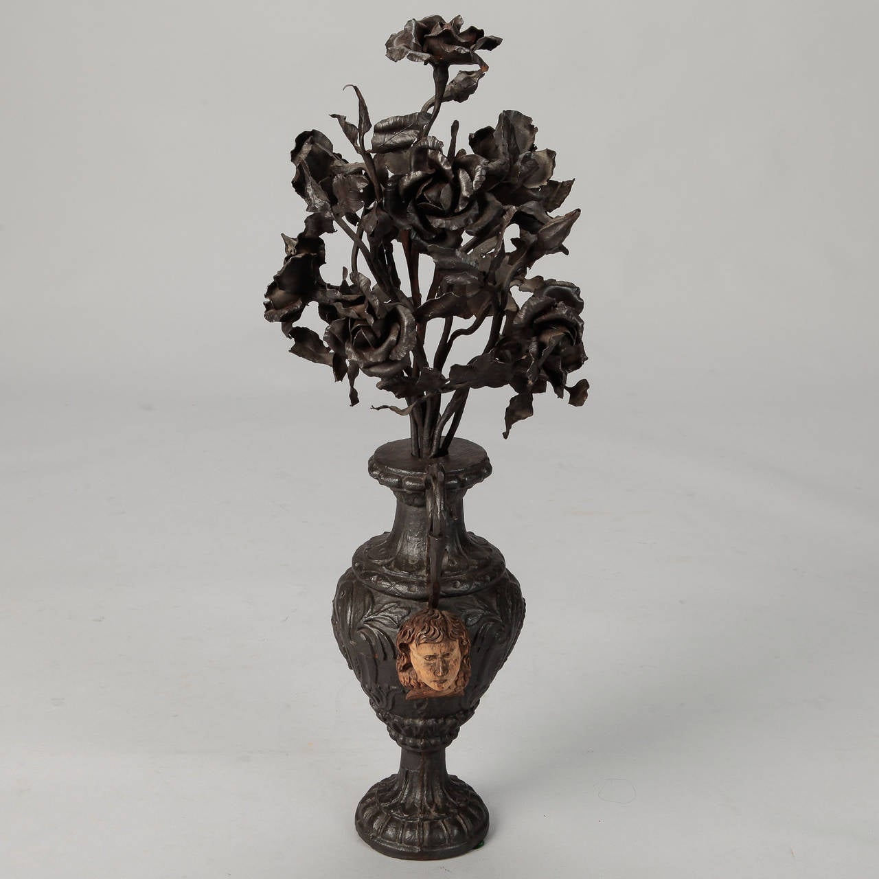 Black cast iron urn is over two feet tall and is topped with intricately rendered roses and decorated with putti faces at the sides, circa 1880s.