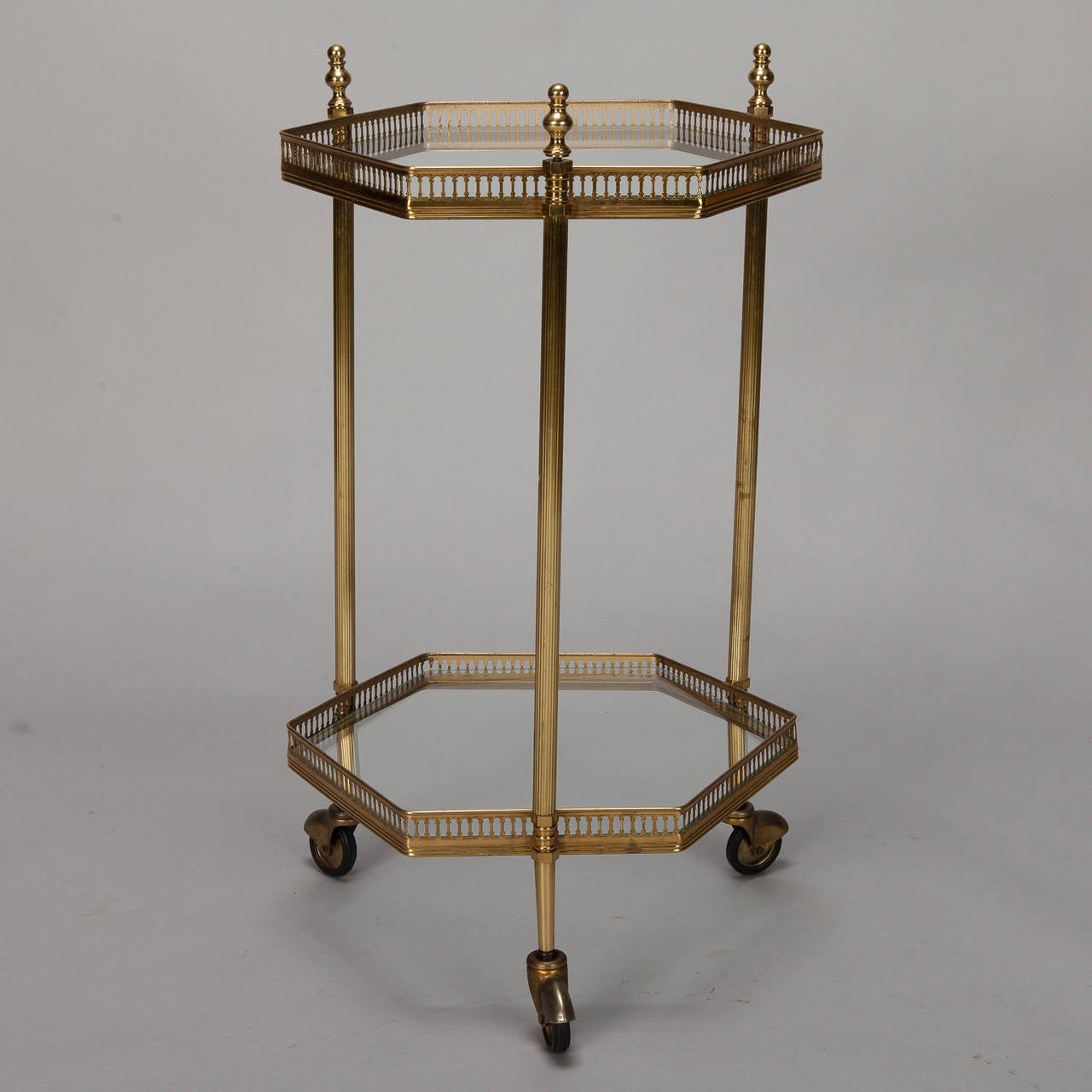 Circa 1960s French brass and glass drinks / serving trolley table has two tiers of six sided glass table tops with open works brass gallery, decorative brass finials topping the three supports and original wheels.