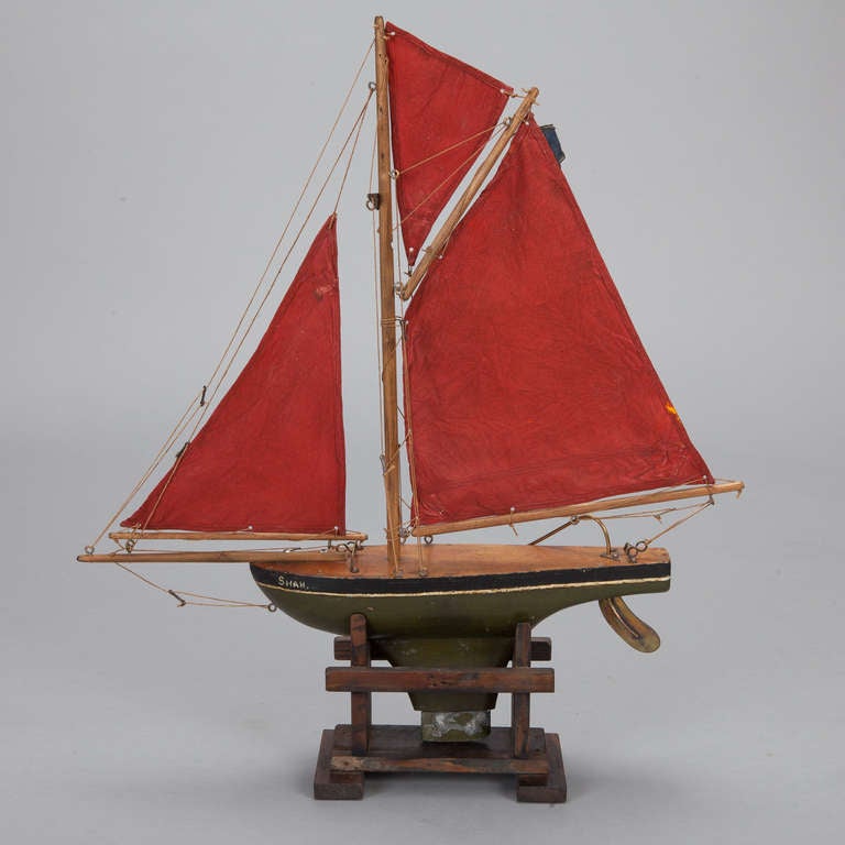 Circa 1900 wooden pond boat has a green rudder, brass fittings, wood deck and three red sails. Display stand shown is included.