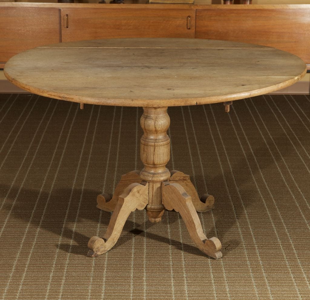 This late 19th century round bleached oak table has a footed pedestal base with carved scrolled feet, a turned column and extendable leaves that fold down.