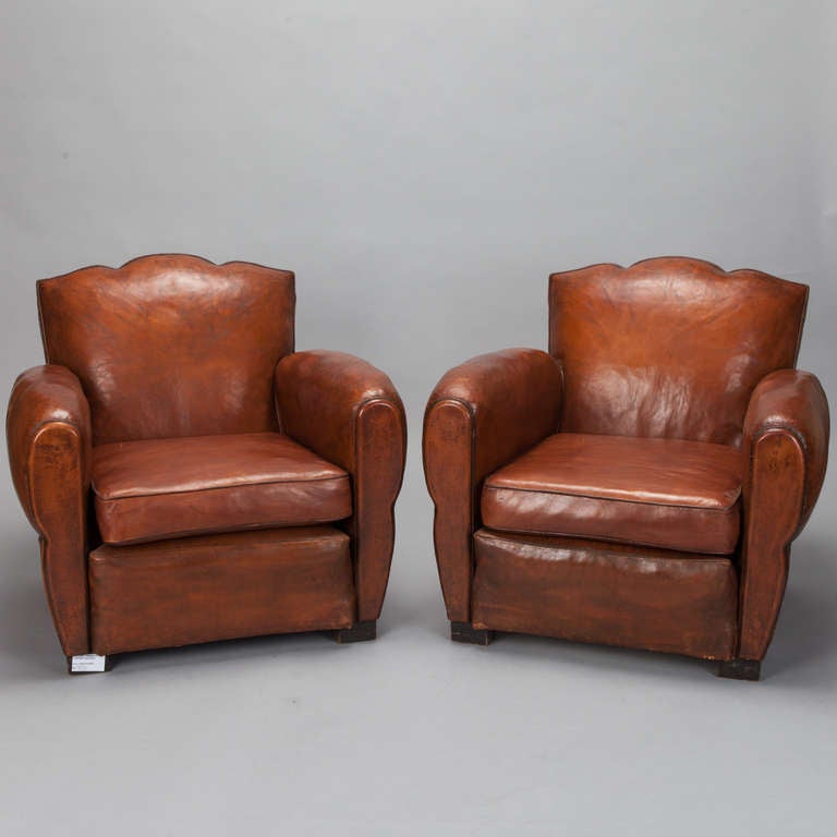 Circa 1930s pair of French club chairs with classic Deco curves, padded arms, wooden feet and upholstered in a rich, medium brown slightly distressed leather.  Chair seat backs are curved with a subtle fluted top. Sold and priced as a pair. 
Arm
