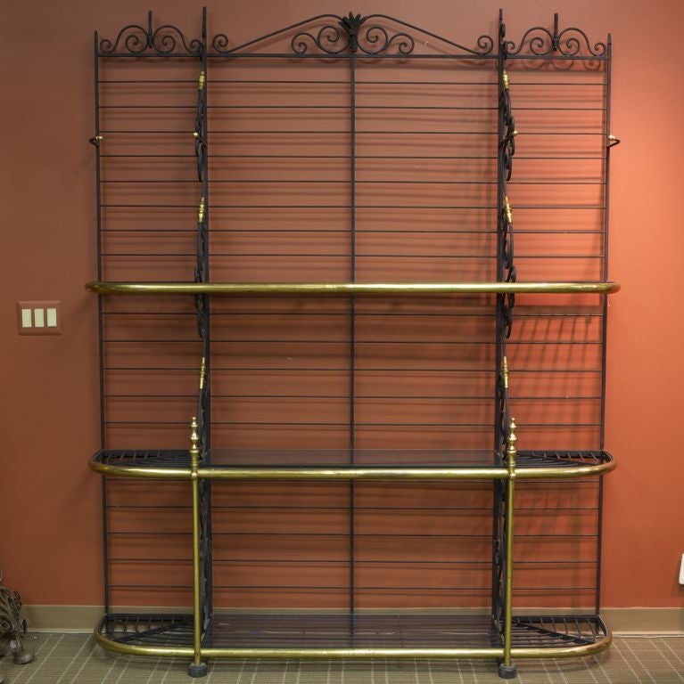 Circa 1930s oversize French bakers rack is made of black iron with decorative scrolled crest, scrolled dividing panels, brass trim and three levels of shelves. The bottom center shelf sections are fitted with glass inserts.