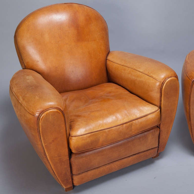 Circa 1930s pair of French leather club chairs with classic Art Deco lines. Rounded seat backs and dramatic, curvy, overstuffed arms upholstered in a warm, caramel colored leather. Seats are 18” high and 21” deep. Arms are 23” high. Sold and priced
