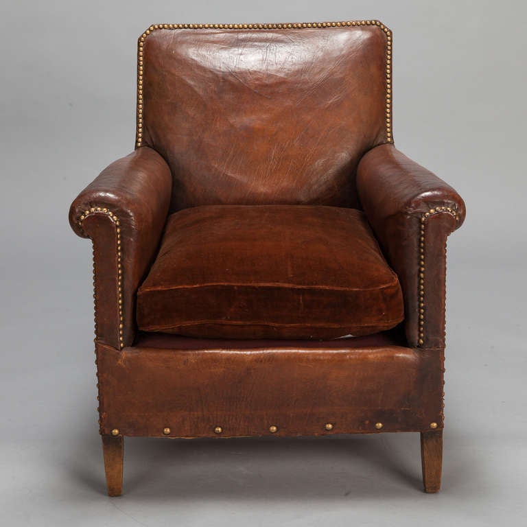 Circa 1930s French Art Deco era armchair in brown leather with rolled arms, brass nailhead trim and a brown velvet seat cushion. Seat is 18” high and 20” deep. Arms are 22.75” high.