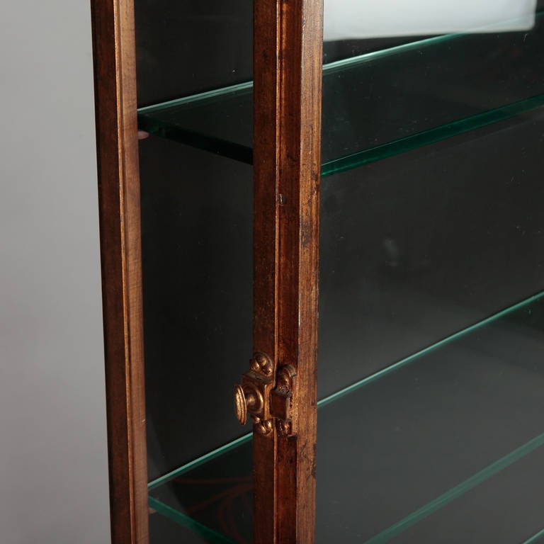 Circa 1960s Spanish shallow metal / iron display cabinet with gilded finish, glass interior shelves and glass doors and sides. This cabinet features delicate tapered legs, hinged side doors and brackets at the top that allow the piece to be bolted