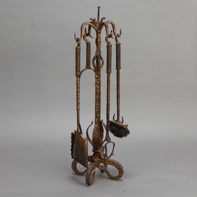 Circa 1940s French fer forge fireplace tool set has a curved, footed base, open work decorative detail on the center support and hooks that support the coordinating tools. Tools include a brush, flat shovel, tongs, and poker. Some heat damage /