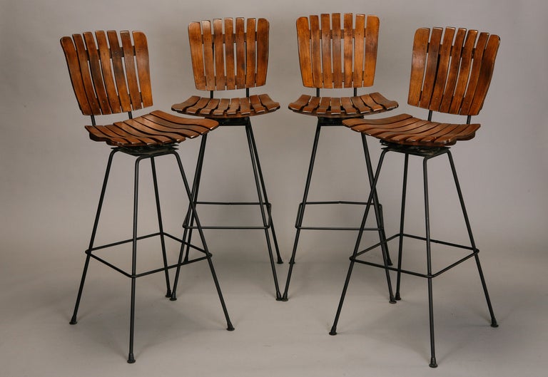 This set of four bar height circa 1950s swivel stools designed by Arthur Umanoff have wrought iron frames with curved slat wood seats and backs. Seats are 30” high from the floor and can be lowered to counter height for an addtional cost.  Sold and