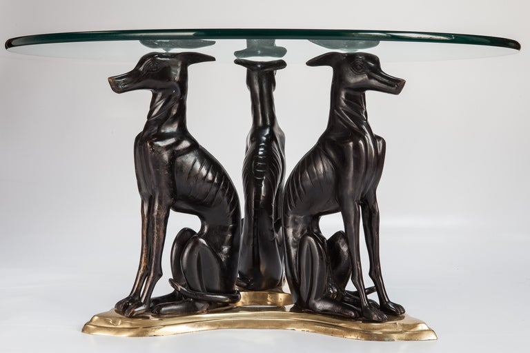 Circa 1980s cocktail table in style of Maitland Smith has a brass base with three cast bronze whippets that support a round glass top. No signature or brand tag found. 

Glass Top:  30” diameter x .5” thick
Base:  19.5” w x 16” h x 30” d
