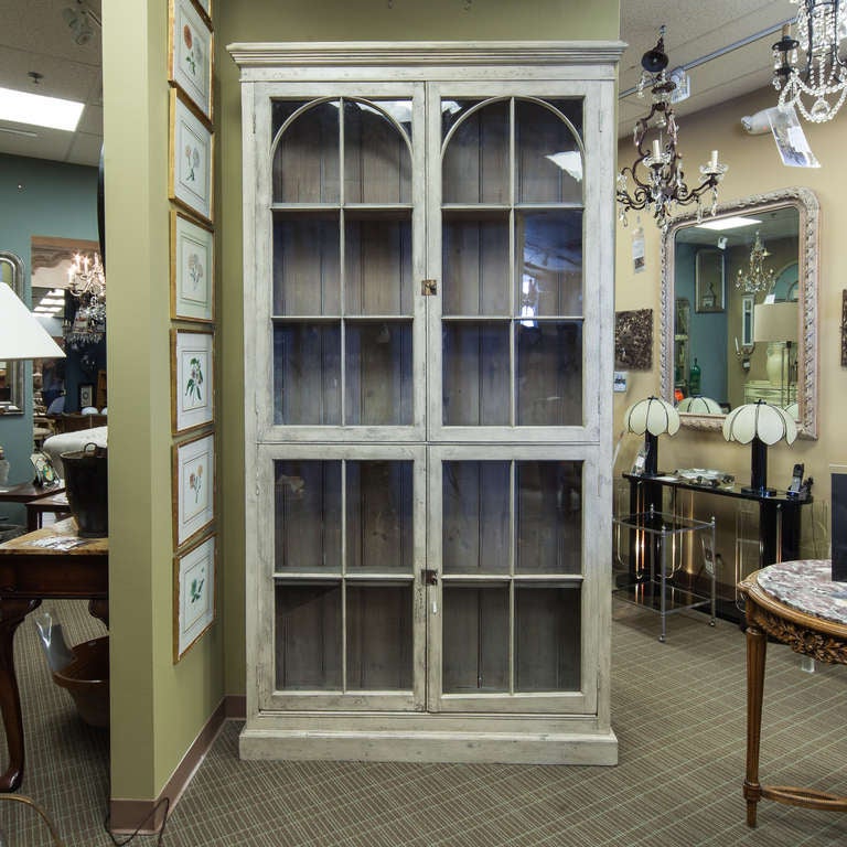 Turn of the century tall French display cabinet in a painted pale gray finish with four hinged glass doors and arched framed details on the top panels. Decorative pediment and shallow depth.