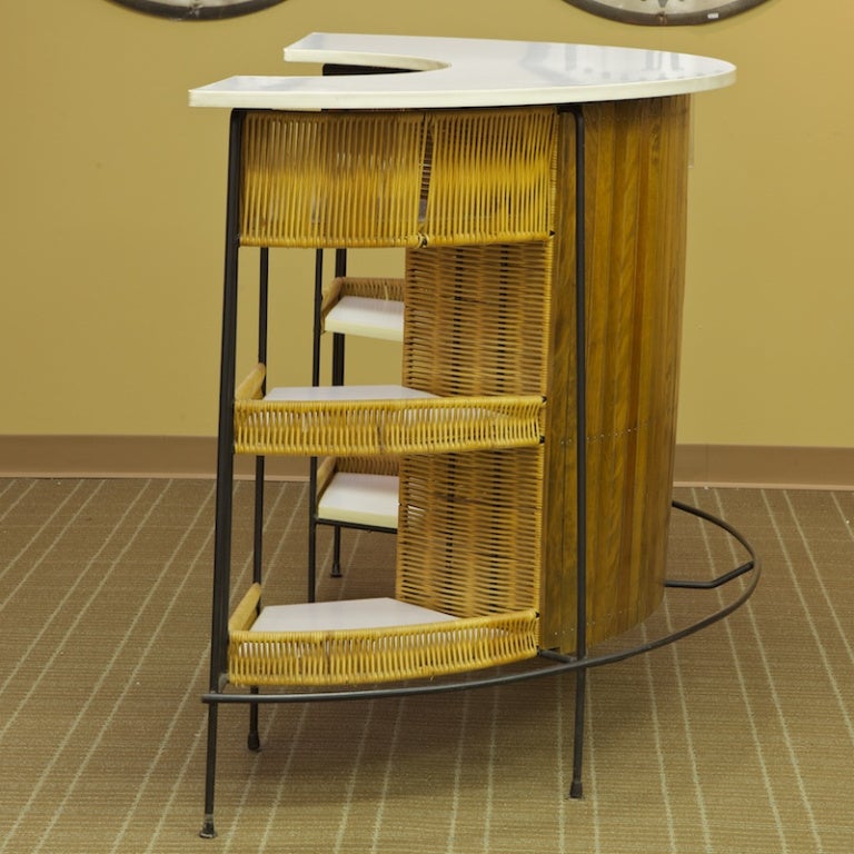 Circa 1950s bar by Arthur Umanoff for Raymor has wrought iron frame, slatted wood front, a white laminate demilune table top and tiered side shelves with rattan accents. This heavy and substantial piece is perfect for entertaining outdoors or in.