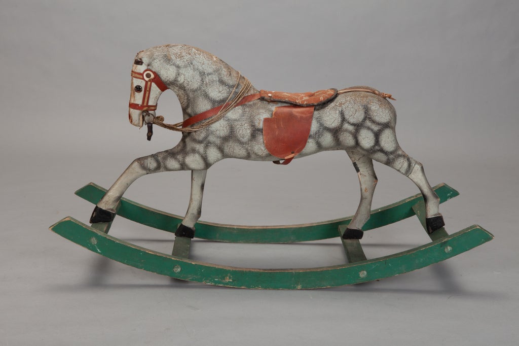 Carved and painted wood rocking horse dates from 1920s-1930s. Horse body is white with gray circles, red bridle and saddle and green rocker. Tail is missing.