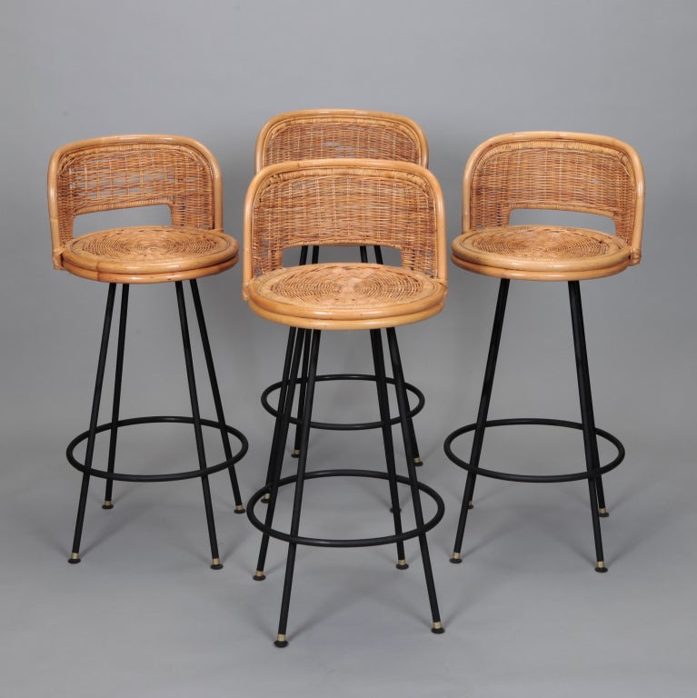 Set of 4 swivel bar stools dated 1966 have black metal four leg base, woven wicker and rattan seats, and are at bar height. In the style of designer Danny Ho Fong. Seats are 28.5” high and 15” deep.