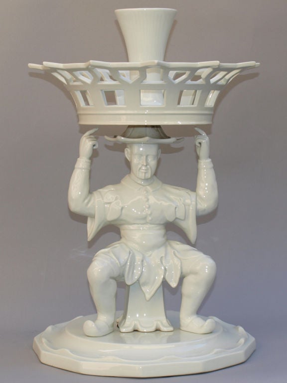 Blanc de Chine epergne, a Chinese male figure in traditional dress, standing with piked legs upon a scalloped plate, a pierced basket cachepot balanced upon his head, centered by a vase. Vista Alegre mark.

Stock ID: D8505.