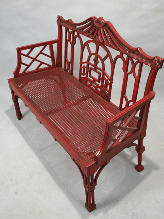 Settee, in Chinese Chippendale taste, having a red lacquered finish, its pierced back shaped as a pagoda roof over arched fretwork, a custom cushion covers its caned seat, raised on column legs.