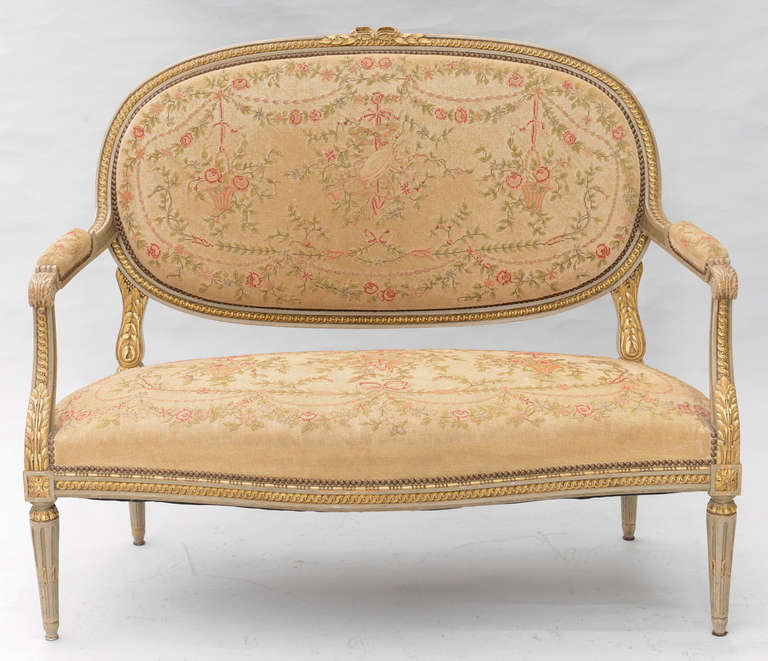 Settee, in Louis XVI style, having a painted and parcel gilt frame, padded medallion backs surmounted by a crest carved as a bow flanked by laureling, joined by acanthine scrolling arms to the padded seat, bowed apron raised on tapering stop-fluted