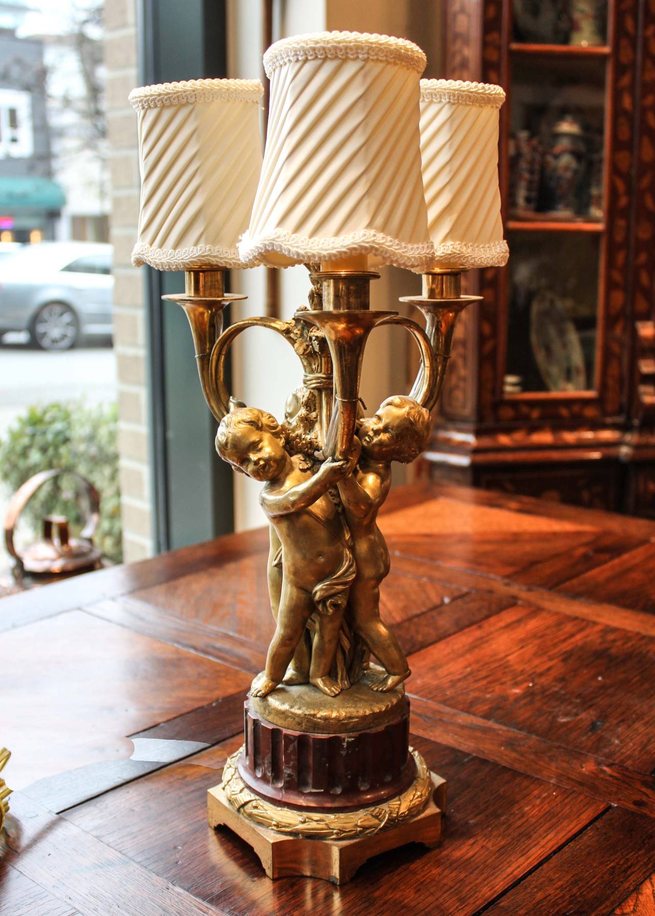 A gilt bronze lamp with beautifully cast cherubic figures supporting three looped candelabra arms. Wired for electricity at a later date, this lamp is otherwise original with a natural patina only attained with age. 

Measures: 5.5